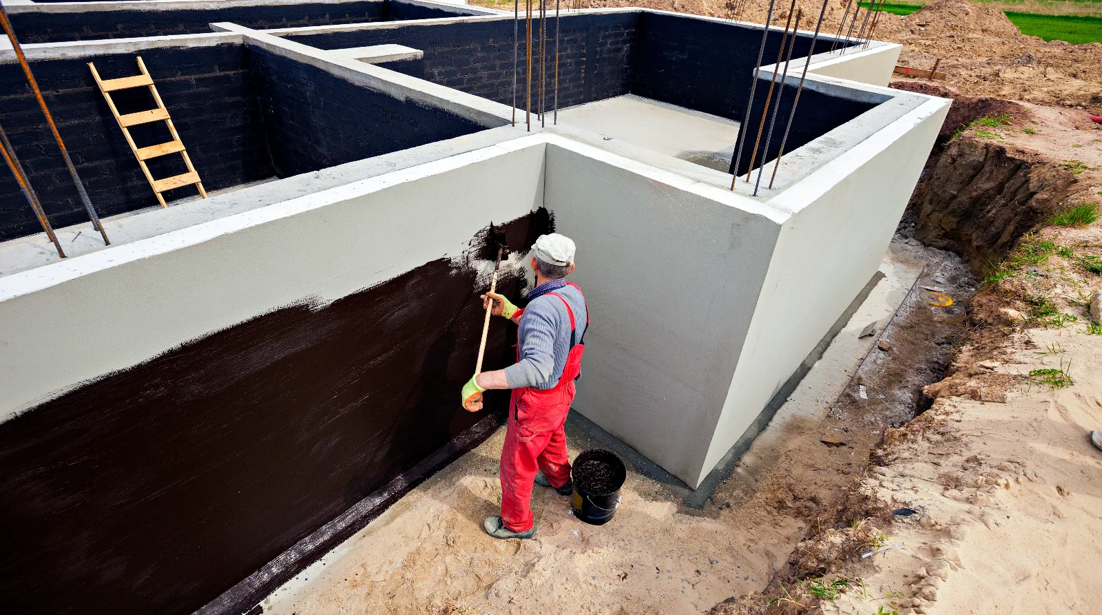 A worker brushing a fluid-applied waterproofing coating onto a concrete foundation wall in an open excavation.